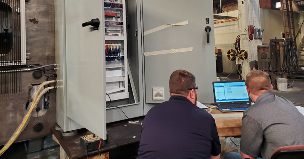 Two techs reviewing data on a laptop beside an open industrial control cabinet in a workshop.